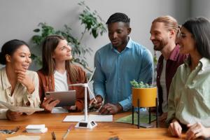 A diverse group of employees engaged in a collaborative meeting around a table, discussing employee evaluation. They are smiling and interacting positively, with plants and a model wind turbine on the table, symbolizing innovation and commitment to core values.